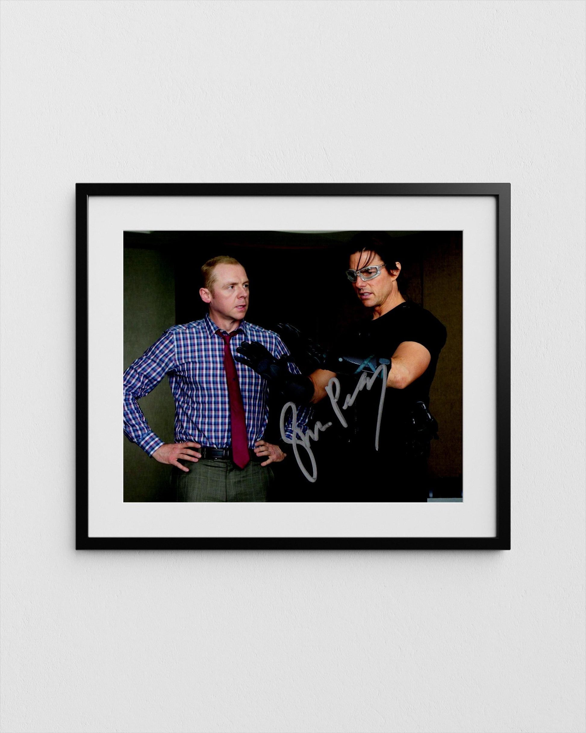 tom cruise and simon pegg, one wearing a blue plaid shirt and the other wearing a black shirt, posing together in a framed photograph.